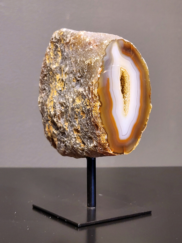 Agaat op standaard, 646 gram, 13cm hg. en 10cm br.  23,- euro, nr. 8
Agaat is een veelzijdige edelsteen die bekend staat om zijn prachtige kleurbanden en patronen. Het is een variëteit van het mineraal kwarts, meer specifiek chalcedoon, en wordt vaak gebruikt in sieraden en als decoratieve steen. 
Algemene kenmerken:
Agaat is een microkristallijne vorm van kwarts, bestaande uit microscopisch kleine kristallen. 
Kleuren en patronen:
Agaat komt voor in vele kleuren, vaak met banden of lagen in verschillende tinten. 
Agaat is een populaire keuze voor het maken van kettingen, armbanden, ringen en andere sieraden vanwege de esthetische aantrekkingskracht en duurzaamheid. 
Spirituele eigenschappen:
Agaat wordt vaak geassocieerd met aardende, stabiliserende en beschermende eigenschappen. 
Helende werking:
Er wordt aangenomen dat agaat kan helpen bij het verminderen van stress, het bevorderen van emotioneel evenwicht en het versterken van het zelfvertrouwen. 
Verzorging van agaat:
Agaat is relatief gemakkelijk te onderhouden. Het kan worden gereinigd met lauw water, de rook van witte salie of palo santo, of door het op een hematiet- of bergkristal-cluster te plaatsen. 

-	Kunstgalerie-Arnold -