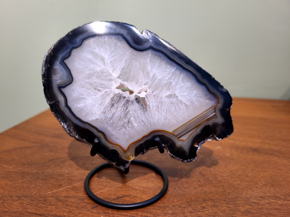 Agaatschijf op standaard, 12cm breed, 13,- euro, nr. 40
Beschrijving Agaat.

Agaat is een veelzijdige edelsteen die bekend staat om zijn prachtige kleurbanden en patronen. Het is een variëteit van het mineraal kwarts, meer specifiek chalcedoon, en wordt vaak gebruikt in sieraden en als decoratieve steen. 
Algemene kenmerken:
Samenstelling:
Agaat is een microkristallijne vorm van kwarts, bestaande uit microscopisch kleine kristallen. 
Kleuren en patronen:
Agaat komt voor in vele kleuren, vaak met banden of lagen in verschillende tinten. 
Hardheid:
Agaat heeft een hardheid van 6,5 tot 7 op de schaal van Mohs, wat betekent dat het een relatief duurzame steen is. 
Herkomst:
De naam "agaat" is afgeleid van de rivier Achates (nu Drillo) op Sicilië, waar de steen voor het eerst werd ontdekt. 
Eigenschappen en toepassingen:
Sieraden:
Agaat is een populaire keuze voor het maken van kettingen, armbanden, ringen en andere sieraden vanwege de esthetische aantrekkingskracht en duurzaamheid. 
Spirituele eigenschappen:
Agaat wordt vaak geassocieerd met aardende, stabiliserende en beschermende eigenschappen. 
Helende werking:
Er wordt aangenomen dat agaat kan helpen bij het verminderen van stress, het bevorderen van emotioneel evenwicht en het versterken van het zelfvertrouwen. 
Andere toepassingen:
Agaat wordt ook gebruikt in decoratieve objecten, zoals schijven en tafels, en werd in het verleden gebruikt in oude culturen voor bescherming en geluk. 
Soorten agaat:
Er zijn veel verschillende soorten agaat, elk met hun eigen unieke kenmerken en eigenschappen, zoals:
Botswana agaat: Bekend om zijn zachte, kalmerende energie en kan helpen bij het loslaten van negatieve emoties. 
Witte agaat: Vaak geassocieerd met innerlijke rust en evenwicht. 
Grijze agaat: Kan helpen bij het stellen van orde en het vinden van stabiliteit. 
Agaat geode: Kan rustgevend werken en chaos tegengaan. 
Verzorging van agaat:
Agaat is relatief gemakkelijk te onderhouden. Het kan worden gereinigd met lauw water, de rook van witte salie of palo santo, of door het op een hematiet- of bergkristal-cluster te plaatsen.