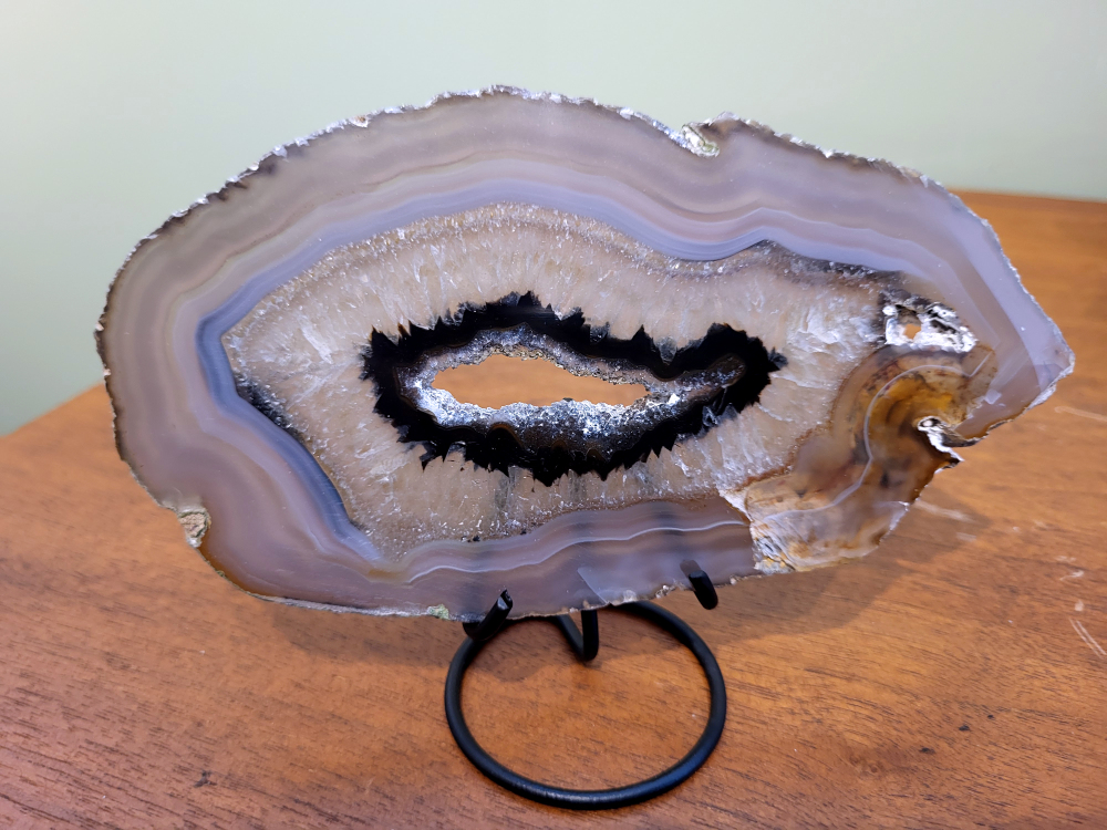 Agaatschijf op standaard 16cm breed, 17,- euro, nr. 43
Beschrijving Agaat.

Agaat is een veelzijdige edelsteen die bekend staat om zijn prachtige kleurbanden en patronen. Het is een variëteit van het mineraal kwarts, meer specifiek chalcedoon, en wordt vaak gebruikt in sieraden en als decoratieve steen. 
Algemene kenmerken:
Samenstelling:
Agaat is een microkristallijne vorm van kwarts, bestaande uit microscopisch kleine kristallen. 
Kleuren en patronen:
Agaat komt voor in vele kleuren, vaak met banden of lagen in verschillende tinten. 
Hardheid:
Agaat heeft een hardheid van 6,5 tot 7 op de schaal van Mohs, wat betekent dat het een relatief duurzame steen is. 
Herkomst:
De naam "agaat" is afgeleid van de rivier Achates (nu Drillo) op Sicilië, waar de steen voor het eerst werd ontdekt. 
Eigenschappen en toepassingen:
Sieraden:
Agaat is een populaire keuze voor het maken van kettingen, armbanden, ringen en andere sieraden vanwege de esthetische aantrekkingskracht en duurzaamheid. 
Spirituele eigenschappen:
Agaat wordt vaak geassocieerd met aardende, stabiliserende en beschermende eigenschappen. 
Helende werking:
Er wordt aangenomen dat agaat kan helpen bij het verminderen van stress, het bevorderen van emotioneel evenwicht en het versterken van het zelfvertrouwen. 
Andere toepassingen:
Agaat wordt ook gebruikt in decoratieve objecten, zoals schijven en tafels, en werd in het verleden gebruikt in oude culturen voor bescherming en geluk. 
Soorten agaat:
Er zijn veel verschillende soorten agaat, elk met hun eigen unieke kenmerken en eigenschappen, zoals:
Botswana agaat: Bekend om zijn zachte, kalmerende energie en kan helpen bij het loslaten van negatieve emoties. 
Witte agaat: Vaak geassocieerd met innerlijke rust en evenwicht. 
Grijze agaat: Kan helpen bij het stellen van orde en het vinden van stabiliteit. 
Agaat geode: Kan rustgevend werken en chaos tegengaan. 
Verzorging van agaat:
Agaat is relatief gemakkelijk te onderhouden. Het kan worden gereinigd met lauw water, de rook van witte salie of palo santo, of door het op een hematiet- of bergkristal-cluster te plaatsen.