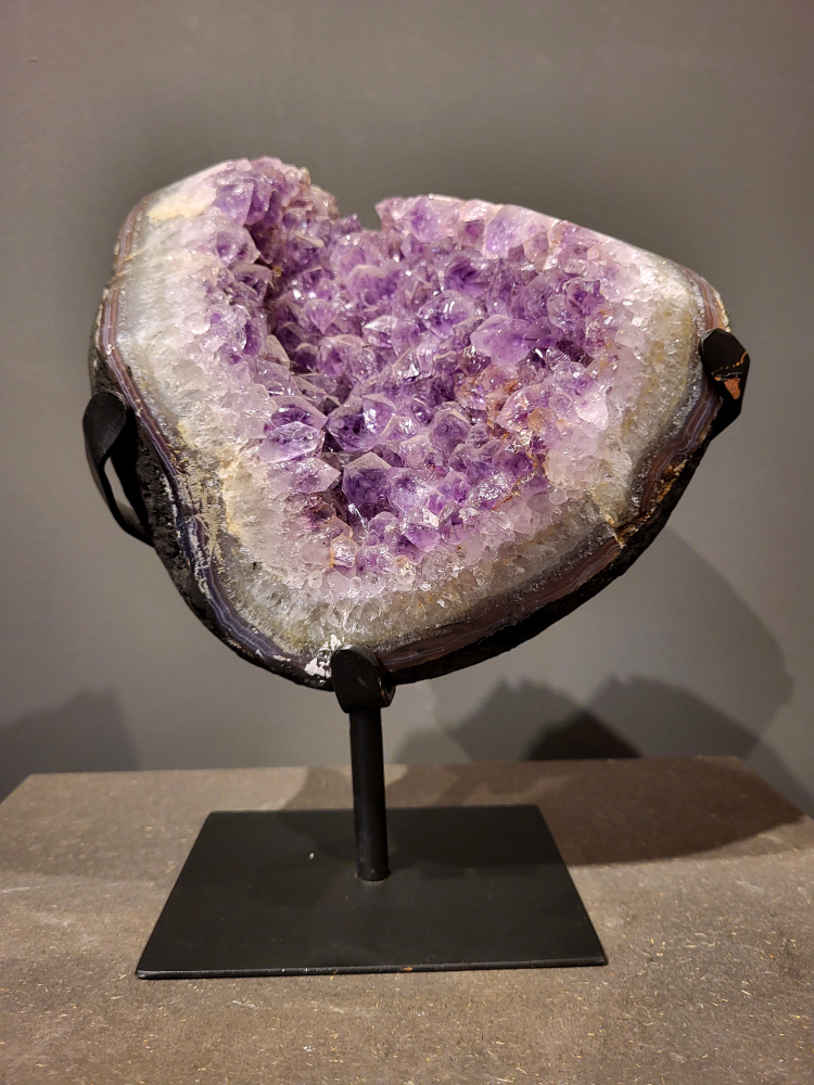 Amethist op standaard, 6,7 kg, 200,- euro, nr. 37 
Waar komt amethist vandaan en hoe ontstaan ze?
Amethist wordt wereldwijd gevonden maar de grootste
vondsten worden gedaan in Brazilië, Uruguay, Mexico en Zambia.
Deze edelsteen komt voor in basalt holtes of geodes die ontstaan zijn door
vulkaanuitbarstingen, het duurt miljoenen jaren voordat de kristallen zich vormen.
De kleur van de amethist wordt veroorzaakt door de aanwezigheid
van ijzer en mangaan. 
Waar kan amethist niet goed tegen?
Amethist kan niet goed tegen fel zonlicht en plotselinge
temperatuurwisselingen. Langdurige blootstelling aan zonlicht
doet de kleur doen vervagen en abrupte temperatuur veranderingen
kan voor barsten en/of scheuren zorgen. Amethist kan daarentegen
wel grotendeels tegen water.
Spirituele werking van de amethist:
> Rust, spirituele groei en bescherming.
Amethist staat vooral bekend om zijn kalmerende werking.

                                                                                        -  Kunstgalerie Arnold  - 