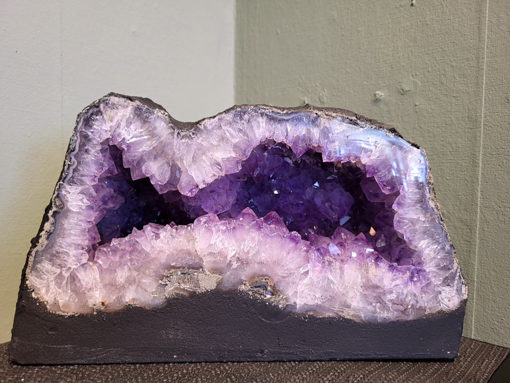 Amethist, 11,28kg, hoogte 18cm, breedte 32cm, 150,- euro, nr. 15
Waar komt amethist vandaan en hoe ontstaan ze?
Amethist wordt wereldwijd gevonden maar de grootste
vondsten worden gedaan in Brazilië, Uruguay, Mexico en Zambia.
Deze edelsteen komt voor in basalt holtes of geodes die ontstaan zijn door
vulkaanuitbarstingen, het duurt miljoenen jaren voordat de kristallen zich vormen.
De kleur van de amethist wordt veroorzaakt door de aanwezigheid
van ijzer en mangaan. 
Waar kan amethist niet goed tegen?
Amethist kan niet goed tegen fel zonlicht en plotselinge
temperatuurwisselingen. Langdurige blootstelling aan zonlicht
doet de kleur doen vervagen en abrupte temperatuur veranderingen
kan voor barsten en/of scheuren zorgen. Amethist kan daarentegen
wel grotendeels tegen water.
Spirituele werking van de amethist:
> Rust, spirituele groei en bescherming.
Amethist staat vooral bekend om zijn kalmerende werking.

                                                                                        -  Kunstgalerie Arnold  - 