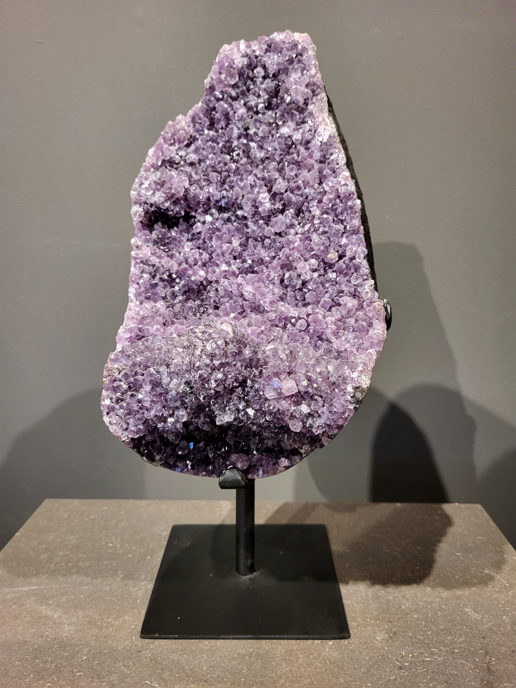 Amethist op standaard, 5,8 kg, 175,- euro, nr. 29 
Waar komt amethist vandaan en hoe ontstaan ze?
Amethist wordt wereldwijd gevonden maar de grootste
vondsten worden gedaan in Brazilië, Uruguay, Mexico en Zambia.
Deze edelsteen komt voor in basalt holtes of geodes die ontstaan zijn door
vulkaanuitbarstingen, het duurt miljoenen jaren voordat de kristallen zich vormen.
De kleur van de amethist wordt veroorzaakt door de aanwezigheid
van ijzer en mangaan. 
Waar kan amethist niet goed tegen?
Amethist kan niet goed tegen fel zonlicht en plotselinge
temperatuurwisselingen. Langdurige blootstelling aan zonlicht
doet de kleur doen vervagen en abrupte temperatuur veranderingen
kan voor barsten en/of scheuren zorgen. Amethist kan daarentegen
wel grotendeels tegen water.
Spirituele werking van de amethist:
> Rust, spirituele groei en bescherming.
Amethist staat vooral bekend om zijn kalmerende werking.

                                                                                        -  Kunstgalerie Arnold  - 