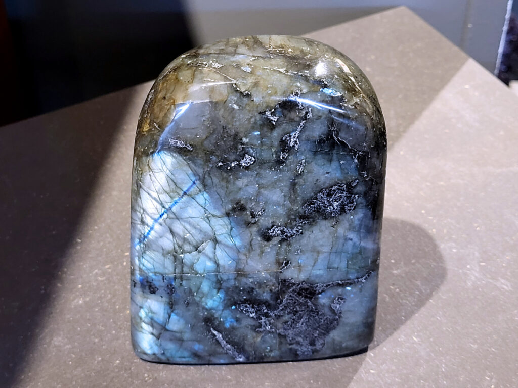 Labradoriet, 1,9kg, 16cm hoog, 57,- euro, nr. 13
Beschrijving, Labradoriet.
Labradoriet is een edelsteen die bekend staat om zijn iriserende kleurenspel, ook wel labradoriseren genoemd, dat vaak doet denken aan het noorderlicht. Het is een populaire steen in de edelstenenwereld vanwege zowel zijn uiterlijk als zijn vermeende spirituele eigenschappen. 
Wat is labradoriet?
Labradoriet is een veldspaatmineraal dat wordt gekenmerkt door een opaalachtig glans of iriserend effect, veroorzaakt door de lichtbreking in de interne structuur van de steen.
De meest voorkomende kleur is grijs of groenachtig grijs, maar de steen kan ook blauwe, gele, oranje, paarse of zelfs rode flitsen laten zien.
De steen is genoemd naar het Canadese schiereiland Labrador, waar het voor het eerst werd ontdekt. 
Spirituele betekenis en eigenschappen:
Labradoriet wordt vaak beschouwd als een beschermende steen, vooral voor mensen die gevoelig zijn voor de energieën van hun omgeving. 
Het zou helpen bij het versterken van de aura en het verminderen van energielekkage, waardoor je je weerbaarder voelt. 
Labradoriet wordt geassocieerd met het versterken van intuïtie, het vergroten van het bewustzijn en het transformeren van negatieve energieën. 
Het wordt ook gebruikt om blokkades op te heffen en de innerlijke wijsheid te onthullen. 
Gebruik:
Labradoriet wordt vaak verwerkt in sieraden zoals hangers, armbanden en oorbellen. 
Het kan ook als ruwe steen, knuffelsteen of in andere vormen worden gebruikt, bijvoorbeeld in een huis of op een werkplek. 
Labradoriet kan worden gereinigd door het onder stromend water te houden of door het te reinigen met een andere steen zoals bergkristal of seleniet. 
Vindplaatsen:
Labradoriet wordt gevonden op verschillende locaties over de hele wereld, waaronder Canada, Finland, Madagaskar en Rusland. 
                                -Kunstgalerie Arnold-