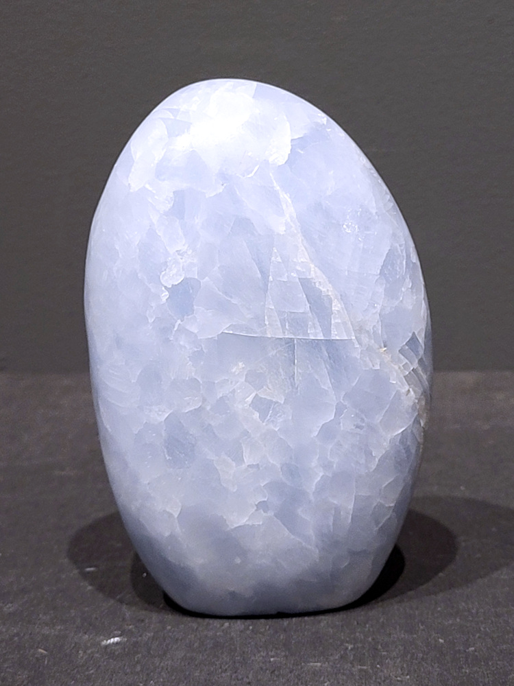 Blauwe calciet, 0,7kg, 12cm hoog, 22,- euro, nr. 20
Calciet is een veelvoorkomend mineraal dat voornamelijk bestaat uit calciumcarbonaat. Het komt in diverse kleuren en vormen voor en wordt in veel industrieën gebruikt, maar ook in de esoterie. 
Algemene kenmerken:
Samenstelling: Calciet bestaat voornamelijk uit calciumcarbonaat (CaCO3). 
Vorming: Het ontstaat door afzetting van calciumcarbonaat in water, maar ook door ophoping van schelpen en skeletten van zeedieren. 
Uiterlijk: Het heeft vaak een glasachtige glans, is doorzichtig tot doorschijnend en kan vele kleuren hebben, zoals geel, oranje, rood, bruin, blauw, groen, roze, wit en kleurloos. 
Hardheid: Calciet heeft een hardheid van 3 op de schaal van Mohs. 
Veelvoorkomend: Het is een van de meest voorkomende mineralen en is een hoofdbestanddeel van kalksteen, marmer en krijt. 
Esoterie:
In de edelsteentherapie wordt calciet gezien als een steen die energie versterkt, negatieve energieën verwijdert en spirituele groei bevordert. Verschillende kleuren calciet hebben specifieke eigenschappen, zoals gele calciet voor doorzettingsvermogen en moed, en mangano calciet voor emotionele heling. 
Specifieke calcietsoorten:
Gele Calciet: Staat bekend om het absorberen van negativiteit, het versterken van doorzettingsvermogen en het aantrekken van succes. 
Blauwe Calciet: Wordt geassocieerd met kalmering, communicatie en het loslaten van stress. 
Groene Calciet: Werkt op de hartchakra, bevordert liefde, compassie en emotionele heling. 
Mangano Calciet: Wordt gebruikt voor emotionele heling, het loslaten van oud verdriet en het versterken van zelfvertrouwen. 
Reiniging:
Calciet kan op verschillende manieren gereinigd worden, maar het is belangrijk om te weten dat sommige soorten, zoals gele calciet, niet goed tegen water kunnen. 
Andere reinigingsmethoden zijn bijvoorbeeld reinigen met edelstenenspray, op een Amethist of bergkristal leggen, of reinigen met selenite. 
Het is een steen die kan worden gebruikt voor energieversterking, emotionele heling en het bevorderen van spirituele groei, afhankelijk van de specifieke kleur en eigenschappen. 
                                                    -Kunstgalerie Arnold-