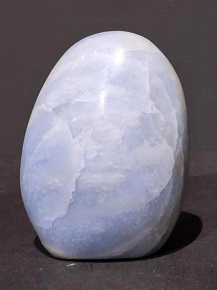 Blauwe calciet, 0,9kg, 11cm hoog, 27,- euro, nr. 26
Calciet is een veelvoorkomend mineraal dat voornamelijk bestaat uit calciumcarbonaat. Het komt in diverse kleuren en vormen voor en wordt in veel industrieën gebruikt, maar ook in de esoterie. 
Algemene kenmerken:
Samenstelling: Calciet bestaat voornamelijk uit calciumcarbonaat (CaCO3). 
Vorming: Het ontstaat door afzetting van calciumcarbonaat in water, maar ook door ophoping van schelpen en skeletten van zeedieren. 
Uiterlijk: Het heeft vaak een glasachtige glans, is doorzichtig tot doorschijnend en kan vele kleuren hebben, zoals geel, oranje, rood, bruin, blauw, groen, roze, wit en kleurloos. 
Hardheid: Calciet heeft een hardheid van 3 op de schaal van Mohs. 
Veelvoorkomend: Het is een van de meest voorkomende mineralen en is een hoofdbestanddeel van kalksteen, marmer en krijt. 
Esoterie:
In de edelsteentherapie wordt calciet gezien als een steen die energie versterkt, negatieve energieën verwijdert en spirituele groei bevordert. Verschillende kleuren calciet hebben specifieke eigenschappen, zoals gele calciet voor doorzettingsvermogen en moed, en mangano calciet voor emotionele heling. 
Specifieke calcietsoorten:
Gele Calciet: Staat bekend om het absorberen van negativiteit, het versterken van doorzettingsvermogen en het aantrekken van succes. 
Blauwe Calciet: Wordt geassocieerd met kalmering, communicatie en het loslaten van stress. 
Groene Calciet: Werkt op de hartchakra, bevordert liefde, compassie en emotionele heling. 
Mangano Calciet: Wordt gebruikt voor emotionele heling, het loslaten van oud verdriet en het versterken van zelfvertrouwen. 
Reiniging:
Calciet kan op verschillende manieren gereinigd worden, maar het is belangrijk om te weten dat sommige soorten, zoals gele calciet, niet goed tegen water kunnen. 
Andere reinigingsmethoden zijn bijvoorbeeld reinigen met edelstenenspray, op een Amethist of bergkristal leggen, of reinigen met selenite. 
Het is een steen die kan worden gebruikt voor energieversterking, emotionele heling en het bevorderen van spirituele groei, afhankelijk van de specifieke kleur en eigenschappen. 
                                                    -Kunstgalerie Arnold-