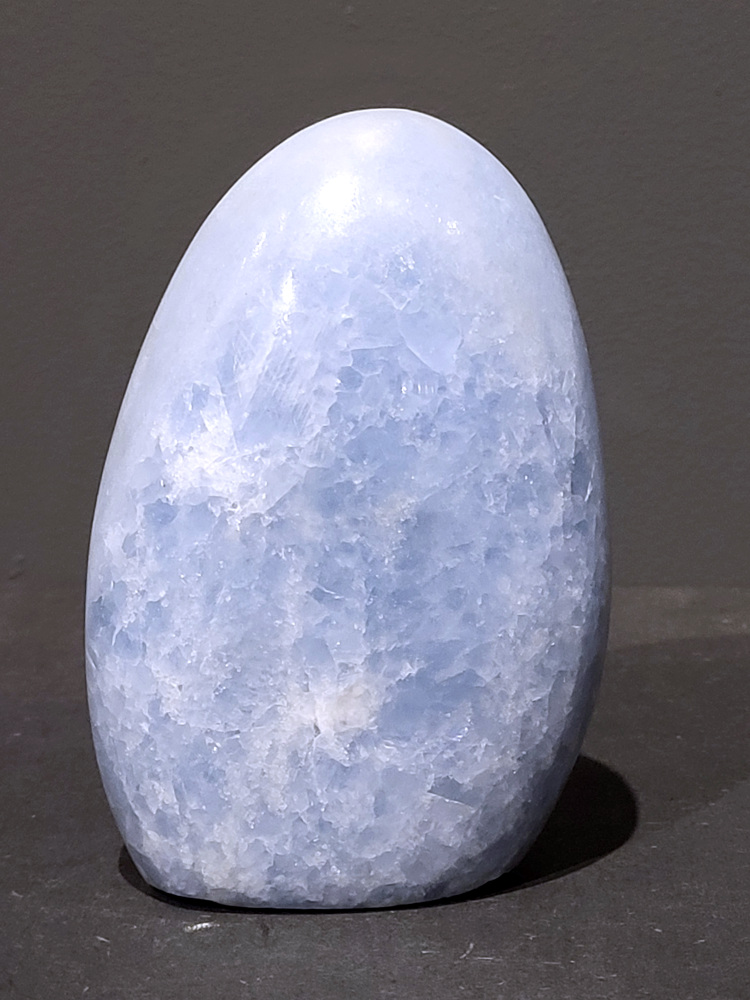 Blauwe calciet, 0,9kg, 12cm hoog, 27,- euro, nr.30
Calciet is een veelvoorkomend mineraal dat voornamelijk bestaat uit calciumcarbonaat. Het komt in diverse kleuren en vormen voor en wordt in veel industrieën gebruikt, maar ook in de esoterie. 
Algemene kenmerken:
Samenstelling: Calciet bestaat voornamelijk uit calciumcarbonaat (CaCO3). 
Vorming: Het ontstaat door afzetting van calciumcarbonaat in water, maar ook door ophoping van schelpen en skeletten van zeedieren. 
Uiterlijk: Het heeft vaak een glasachtige glans, is doorzichtig tot doorschijnend en kan vele kleuren hebben, zoals geel, oranje, rood, bruin, blauw, groen, roze, wit en kleurloos. 
Hardheid: Calciet heeft een hardheid van 3 op de schaal van Mohs. 
Veelvoorkomend: Het is een van de meest voorkomende mineralen en is een hoofdbestanddeel van kalksteen, marmer en krijt. 
Esoterie:
In de edelsteentherapie wordt calciet gezien als een steen die energie versterkt, negatieve energieën verwijdert en spirituele groei bevordert. Verschillende kleuren calciet hebben specifieke eigenschappen, zoals gele calciet voor doorzettingsvermogen en moed, en mangano calciet voor emotionele heling. 
Specifieke calcietsoorten:
Gele Calciet: Staat bekend om het absorberen van negativiteit, het versterken van doorzettingsvermogen en het aantrekken van succes. 
Blauwe Calciet: Wordt geassocieerd met kalmering, communicatie en het loslaten van stress. 
Groene Calciet: Werkt op de hartchakra, bevordert liefde, compassie en emotionele heling. 
Mangano Calciet: Wordt gebruikt voor emotionele heling, het loslaten van oud verdriet en het versterken van zelfvertrouwen. 
Reiniging:
Calciet kan op verschillende manieren gereinigd worden, maar het is belangrijk om te weten dat sommige soorten, zoals gele calciet, niet goed tegen water kunnen. 
Andere reinigingsmethoden zijn bijvoorbeeld reinigen met edelstenenspray, op een Amethist of bergkristal leggen, of reinigen met selenite. 
Het is een steen die kan worden gebruikt voor energieversterking, emotionele heling en het bevorderen van spirituele groei, afhankelijk van de specifieke kleur en eigenschappen. 
                                                    -Kunstgalerie Arnold-