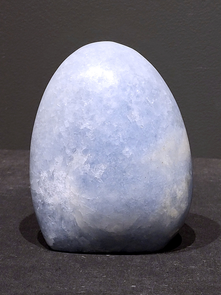 blauwe calciet, 0,8kg, 12cm hoog, 25,- euro, nr. 25
Calciet is een veelvoorkomend mineraal dat voornamelijk bestaat uit calciumcarbonaat. Het komt in diverse kleuren en vormen voor en wordt in veel industrieën gebruikt, maar ook in de esoterie. 
Algemene kenmerken:
Samenstelling: Calciet bestaat voornamelijk uit calciumcarbonaat (CaCO3). 
Vorming: Het ontstaat door afzetting van calciumcarbonaat in water, maar ook door ophoping van schelpen en skeletten van zeedieren. 
Uiterlijk: Het heeft vaak een glasachtige glans, is doorzichtig tot doorschijnend en kan vele kleuren hebben, zoals geel, oranje, rood, bruin, blauw, groen, roze, wit en kleurloos. 
Hardheid: Calciet heeft een hardheid van 3 op de schaal van Mohs. 
Veelvoorkomend: Het is een van de meest voorkomende mineralen en is een hoofdbestanddeel van kalksteen, marmer en krijt. 
Esoterie:
In de edelsteentherapie wordt calciet gezien als een steen die energie versterkt, negatieve energieën verwijdert en spirituele groei bevordert. Verschillende kleuren calciet hebben specifieke eigenschappen, zoals gele calciet voor doorzettingsvermogen en moed, en mangano calciet voor emotionele heling. 
Specifieke calcietsoorten:
Gele Calciet: Staat bekend om het absorberen van negativiteit, het versterken van doorzettingsvermogen en het aantrekken van succes. 
Blauwe Calciet: Wordt geassocieerd met kalmering, communicatie en het loslaten van stress. 
Groene Calciet: Werkt op de hartchakra, bevordert liefde, compassie en emotionele heling. 
Mangano Calciet: Wordt gebruikt voor emotionele heling, het loslaten van oud verdriet en het versterken van zelfvertrouwen. 
Reiniging:
Calciet kan op verschillende manieren gereinigd worden, maar het is belangrijk om te weten dat sommige soorten, zoals gele calciet, niet goed tegen water kunnen. 
Andere reinigingsmethoden zijn bijvoorbeeld reinigen met edelstenenspray, op een Amethist of bergkristal leggen, of reinigen met selenite. 
Het is een steen die kan worden gebruikt voor energieversterking, emotionele heling en het bevorderen van spirituele groei, afhankelijk van de specifieke kleur en eigenschappen. 
                                                    -Kunstgalerie Arnold-