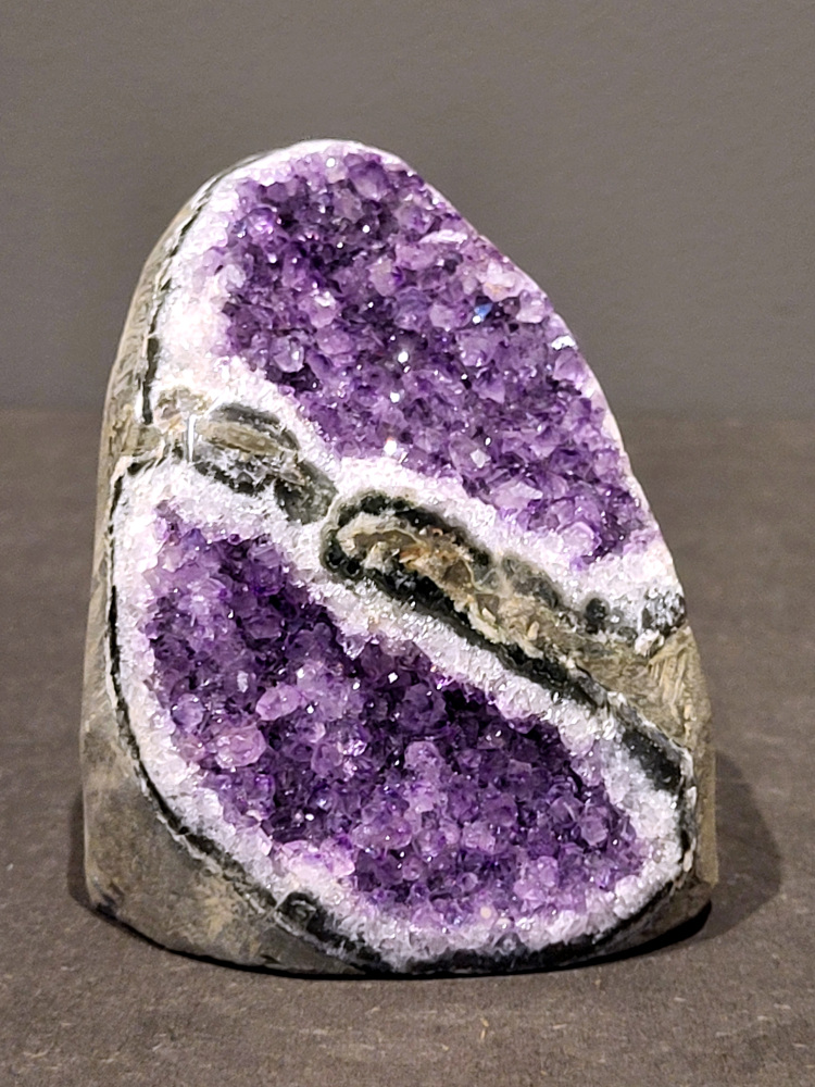 Amethist druzy, 0,8kg, 34,- euro, nr. 39

Beschrijving Amethist.
Amethist is een paarse variëteit van kwarts, bekend om zijn kalmerende en spirituele eigenschappen. De kleur varieert van licht lila tot diep violet en wordt veroorzaakt door sporen van ijzer en andere elementen. Amethist wordt al eeuwenlang gewaardeerd, zowel vanwege zijn esthetische schoonheid als zijn vermeende genezende en beschermende krachten. 
Amethist is een kwartskristal, vaak te vinden in geodes (holtes in gesteente) of als afzonderlijke kristallen. 
De kleur varieert van lichtpaars tot diep violet, soms met roze of blauwe tinten. 
De kleurintensiteit kan variëren, waarbij donkerdere amethisten vaak als kwalitatief worden beschouwd. 
Soms vertoont amethist dichroïsme, waarbij je verschillende kleuren kunt zien wanneer je de steen in het licht draait. 

Vindplaatsen:
Amethist wordt gevonden in verschillende delen van de wereld, waaronder Brazilië, Uruguay, Zambia, Madagaskar, Rusland en Canada.
Het wordt vaak gevonden in bergachtige gebieden. 

Gebruik:
Amethist wordt gebruikt in sieraden, decoratieve objecten en als helende steen. 
Het wordt vaak gedragen als hanger, ring of armband, of geplaatst in ruimtes om de energie te zuiveren. 
Amethist wordt ook gebruikt in meditatie en andere spirituele praktijken. 