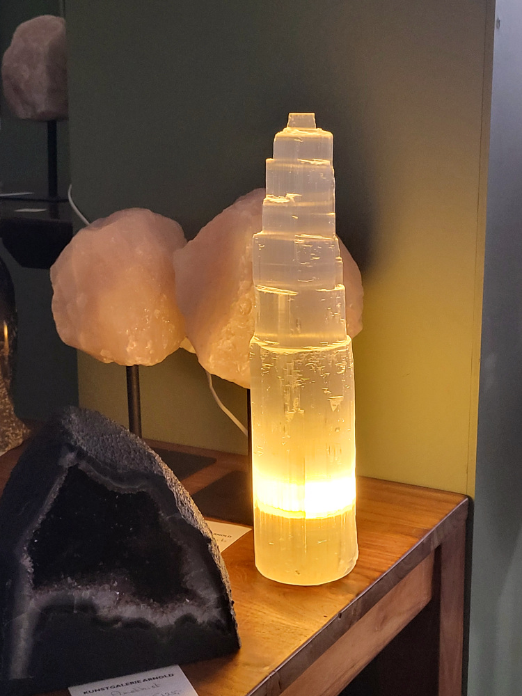 Seleniet lamp, 46cm hoog, XXL formaat, 95,- euro, nr. 53

Seleniet is een kristallijne vorm van gips, ook wel satijnspaat genoemd, bekend om zijn zuiverende en spirituele eigenschappen. Het mineraal is relatief zacht, is te vinden in verschillende kleuren (wit, oranje, geel, groen) en wordt vaak gebruikt bij healing en meditatie. Seleniet wordt spiritueel geassocieerd met rust, heling en het reinigen van energie, en het staat bekend om zijn zelfreinigende en het reinigen van andere stenen. 
Eigenschappen en uiterlijk
Samenstelling: Seleniet is een gehydrateerd calciumsulfaat en een variant van gips.
Hardheid: Met een hardheid van 2 is het een zachte steen.
Vorm: Het komt vaak voor in lange, heldere, tabulaire kristallen met een vezelige structuur.
Kleur: De kleur kan variëren, maar de meest voorkomende kleuren zijn wit, kleurloos, geel, oranje en groen. De witte variant wordt vaak satijnspaat genoemd.
Bijzonderheden: Sommige selenietkristallen kunnen flexibel zijn en enhydrokristallen kunnen kleine water- en gasbelletjes bevatten. 
Spirituele betekenis en gebruik
Reiniging: Seleniet wordt beschouwd als een krachtige zuiveraar die negatieve energie verwijdert en harmonie brengt.
Healing en meditatie: Het wordt vaak gebruikt tijdens meditatie, healing en voor het communiceren met hogere sferen.
Zelfreinigend: Seleniet is een zelfreinigende steen en wordt vaak gebruikt om andere edelstenen energetisch op te laden.
Chakra's: Het is sterk verbonden met de kruin- en derde-oogchakra, wat helpt bij spiritueel bewustzijn en intuïtie.
Spirituele namen: Het wordt ook wel "engelensteen" genoemd vanwege de hoge vibratie die heling en transformatie brengt. 
Verzorging
Vermijd water: Omdat het een gipsvariant is, mag seleniet nooit worden gereinigd met water, aangezien het kan oplossen of beschadigen.
Reinigingsmethodes: Reinig het energetisch met rook van salie of palo santo, of door het op een cluster van amethist of hematiet te plaatsen.
Opladen: Om seleniet op te laden, kan maanlicht of een bergkristal worden gebruikt.