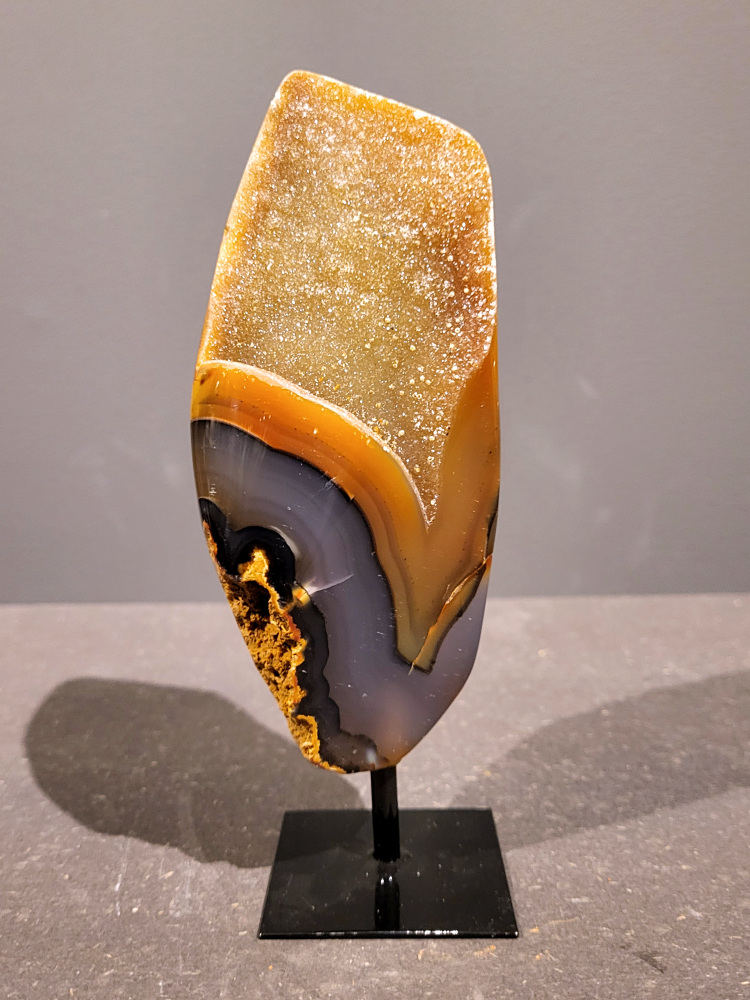 <strong>Agaat op standaard, 0,7 kg, 19cm hoog, 35,- euro, nr. 32</strong>

<strong>Beschrijving Agaat.</strong>

Agaat is een veelzijdige edelsteen die bekend staat om zijn prachtige kleurbanden en patronen. Het is een variëteit van het mineraal kwarts, meer specifiek chalcedoon, en wordt vaak gebruikt in sieraden en als decoratieve steen. 
<strong>Algemene kenmerken:</strong>
Agaat is een microkristallijne vorm van kwarts, bestaande uit microscopisch kleine kristallen. 
<strong>Kleuren en patronen:</strong>
Agaat komt voor in vele kleuren, vaak met banden of lagen in verschillende tinten. 
Agaat is een populaire keuze voor het maken van kettingen, armbanden, ringen en andere sieraden vanwege de esthetische aantrekkingskracht en duurzaamheid. 
<strong>Spirituele eigenschappen:</strong>
Agaat wordt vaak geassocieerd met aardende, stabiliserende en beschermende eigenschappen. 
<strong>Helende werking:</strong>
Er wordt aangenomen dat agaat kan helpen bij het verminderen van stress, het bevorderen van emotioneel evenwicht en het versterken van het zelfvertrouwen. 
<strong>Verzorging van agaat:</strong>
Agaat is relatief gemakkelijk te onderhouden. Het kan worden gereinigd met lauw water, de rook van witte salie of palo santo, of door het op een hematiet- of bergkristal-cluster te plaatsen. 

<strong>-	Kunstgalerie-Arnold -</strong>