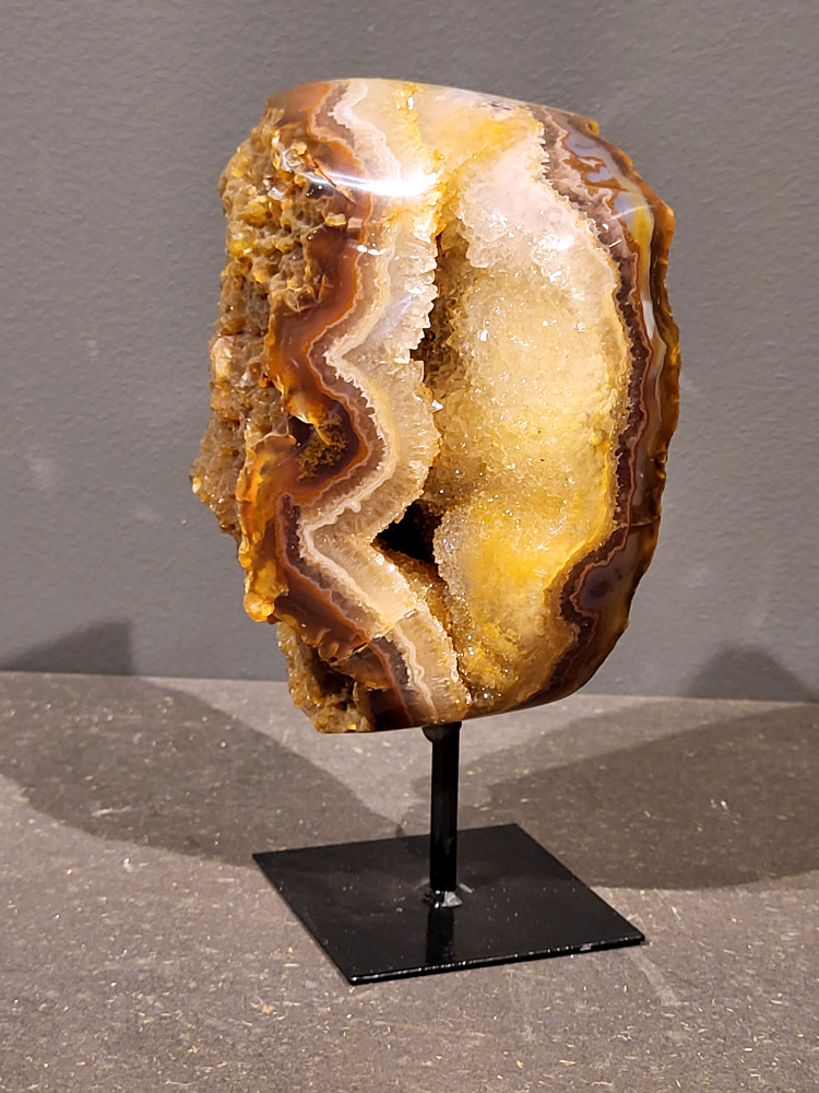 <strong>Agaat op standaard, 19cm hoog, 1,4kg, 71,- euro, nr. 23</strong>

Agaat is een veelzijdige edelsteen die bekend staat om zijn prachtige kleurbanden en patronen. Het is een variëteit van het mineraal kwarts, meer specifiek chalcedoon, en wordt vaak gebruikt in sieraden en als decoratieve steen. 
<strong>Algemene kenmerken:</strong>
Agaat is een microkristallijne vorm van kwarts, bestaande uit microscopisch kleine kristallen. 
<strong>Kleuren en patronen:</strong>
Agaat komt voor in vele kleuren, vaak met banden of lagen in verschillende tinten. 
Agaat is een populaire keuze voor het maken van kettingen, armbanden, ringen en andere sieraden vanwege de esthetische aantrekkingskracht en duurzaamheid. 
<strong>Spirituele eigenschappen:</strong>
Agaat wordt vaak geassocieerd met aardende, stabiliserende en beschermende eigenschappen. 
<strong>Helende werking:</strong>
Er wordt aangenomen dat agaat kan helpen bij het verminderen van stress, het bevorderen van emotioneel evenwicht en het versterken van het zelfvertrouwen. 
<strong>Verzorging van agaat:</strong>
Agaat is relatief gemakkelijk te onderhouden. Het kan worden gereinigd met lauw water, de rook van witte salie of palo santo, of door het op een hematiet- of bergkristal-cluster te plaatsen. 

<strong>-	Kunstgalerie-Arnold -</strong>