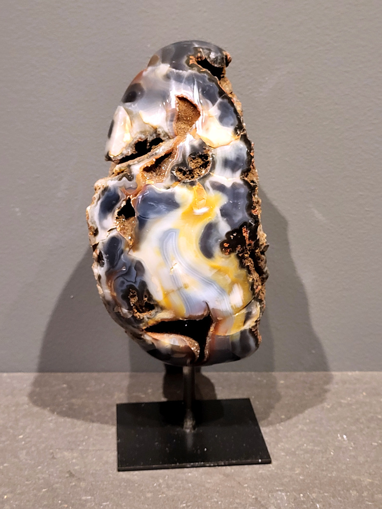 <strong>Agaat op standaard, 21cm hoog, 2 kg, 98,- euro, nr. 18</strong>

Agaat is een veelzijdige edelsteen die bekend staat om zijn prachtige kleurbanden en patronen. Het is een variëteit van het mineraal kwarts, meer specifiek chalcedoon, en wordt vaak gebruikt in sieraden en als decoratieve steen. 
<strong>Algemene kenmerken:</strong>
Agaat is een microkristallijne vorm van kwarts, bestaande uit microscopisch kleine kristallen. 
<strong>Kleuren en patronen:</strong>
Agaat komt voor in vele kleuren, vaak met banden of lagen in verschillende tinten. 
Agaat is een populaire keuze voor het maken van kettingen, armbanden, ringen en andere sieraden vanwege de esthetische aantrekkingskracht en duurzaamheid. 
<strong>Spirituele eigenschappen:</strong>
Agaat wordt vaak geassocieerd met aardende, stabiliserende en beschermende eigenschappen. 
<strong>Helende werking:</strong>
Er wordt aangenomen dat agaat kan helpen bij het verminderen van stress, het bevorderen van emotioneel evenwicht en het versterken van het zelfvertrouwen. 
<strong>Verzorging van agaat:</strong>
Agaat is relatief gemakkelijk te onderhouden. Het kan worden gereinigd met lauw water, de rook van witte salie of palo santo, of door het op een hematiet- of bergkristal-cluster te plaatsen. 

<strong>-	Kunstgalerie-Arnold -</strong>