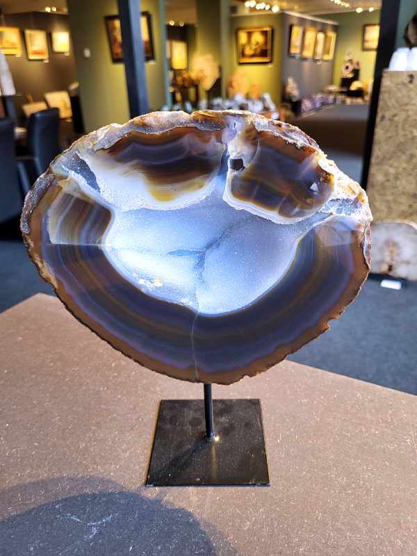 <strong>Agaat op standaard, 3 kg, 149,- euro, nr. 55</strong>

<strong>Agaat is een veelzijdige edelsteen die bekend staat om zijn prachtige kleurbanden en patronen.</strong> Het is een variëteit van het mineraal kwarts, meer specifiek chalcedoon, en wordt vaak gebruikt in sieraden en als decoratieve steen. 
<strong>Algemene kenmerken:</strong>
Agaat is een microkristallijne vorm van kwarts, bestaande uit microscopisch kleine kristallen. 
<strong>Kleuren en patronen:</strong>
Agaat komt voor in vele kleuren, vaak met banden of lagen in verschillende tinten. 
Agaat is een populaire keuze voor het maken van kettingen, armbanden, ringen en andere sieraden vanwege de esthetische aantrekkingskracht en duurzaamheid. 
<strong>Spirituele eigenschappen:</strong>
Agaat wordt vaak geassocieerd met aardende, stabiliserende en beschermende eigenschappen. 
<strong>Helende werking:</strong>
Er wordt aangenomen dat agaat kan helpen bij het verminderen van stress, het bevorderen van emotioneel evenwicht en het versterken van het zelfvertrouwen. 
<strong>Verzorging van agaat:</strong>
Agaat is relatief gemakkelijk te onderhouden. Het kan worden gereinigd met lauw water, de rook van witte salie of palo santo, of door het op een hematiet- of bergkristal-cluster te plaatsen.