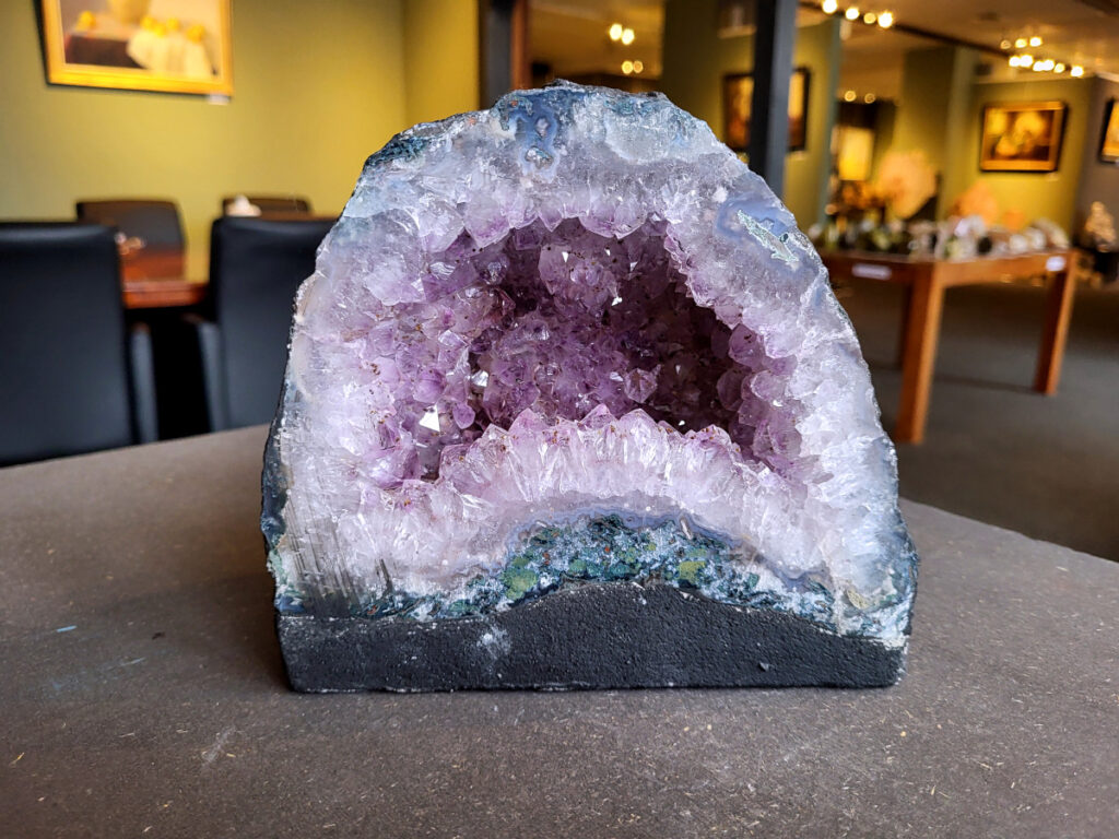 Amethist, Amethist geode met standvlak, Kunstgalerie Arnold, 3,3 kg, 35,- euro, nr. 2

Beschrijving Amethist.
Amethist is een paarse variëteit van kwarts, bekend om zijn kalmerende en spirituele eigenschappen. De kleur varieert van licht lila tot diep violet en wordt veroorzaakt door sporen van ijzer en andere elementen. Amethist wordt al eeuwenlang gewaardeerd, zowel vanwege zijn esthetische schoonheid als zijn vermeende genezende en beschermende krachten. 
Amethist is een kwartskristal, vaak te vinden in geodes (holtes in gesteente) of als afzonderlijke kristallen. 
De kleur varieert van lichtpaars tot diep violet, soms met roze of blauwe tinten. 
De kleurintensiteit kan variëren, waarbij donkerdere amethisten vaak als kwalitatief worden beschouwd. 
Soms vertoont amethist dichroïsme, waarbij je verschillende kleuren kunt zien wanneer je de steen in het licht draait. 

Vindplaatsen:
Amethist wordt gevonden in verschillende delen van de wereld, waaronder Brazilië, Uruguay, Zambia, Madagaskar, Rusland en Canada.
Het wordt vaak gevonden in bergachtige gebieden. 

Gebruik:
Amethist wordt gebruikt in sieraden, decoratieve objecten en als helende steen. 
Het wordt vaak gedragen als hanger, ring of armband, of geplaatst in ruimtes om de energie te zuiveren. 
Amethist wordt ook gebruikt in meditatie en andere spirituele praktijken. 
