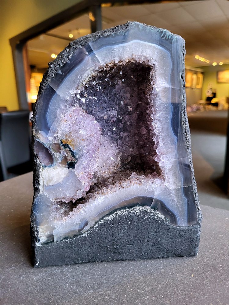 Amethist, Amethist geode met standvlak, Kunstgalerie Arnold, 4,1 kg, 45,- euro, nr. 3

Beschrijving Amethist.
Amethist is een paarse variëteit van kwarts, bekend om zijn kalmerende en spirituele eigenschappen. De kleur varieert van licht lila tot diep violet en wordt veroorzaakt door sporen van ijzer en andere elementen. Amethist wordt al eeuwenlang gewaardeerd, zowel vanwege zijn esthetische schoonheid als zijn vermeende genezende en beschermende krachten. 
Amethist is een kwartskristal, vaak te vinden in geodes (holtes in gesteente) of als afzonderlijke kristallen. 
De kleur varieert van lichtpaars tot diep violet, soms met roze of blauwe tinten. 
De kleurintensiteit kan variëren, waarbij donkerdere amethisten vaak als kwalitatief worden beschouwd. 
Soms vertoont amethist dichroïsme, waarbij je verschillende kleuren kunt zien wanneer je de steen in het licht draait. 

Vindplaatsen:
Amethist wordt gevonden in verschillende delen van de wereld, waaronder Brazilië, Uruguay, Zambia, Madagaskar, Rusland en Canada.
Het wordt vaak gevonden in bergachtige gebieden. 

Gebruik:
Amethist wordt gebruikt in sieraden, decoratieve objecten en als helende steen. 
Het wordt vaak gedragen als hanger, ring of armband, of geplaatst in ruimtes om de energie te zuiveren. 
Amethist wordt ook gebruikt in meditatie en andere spirituele praktijken. 
