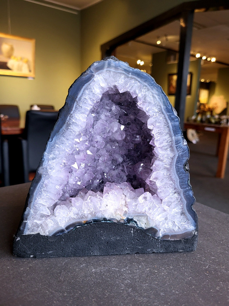 Amethist, Amethist geode met standvlak, Kunstgalerie Arnold, 4,2 kg, 42,- euro, nr. 4

Beschrijving Amethist.
Amethist is een paarse variëteit van kwarts, bekend om zijn kalmerende en spirituele eigenschappen. De kleur varieert van licht lila tot diep violet en wordt veroorzaakt door sporen van ijzer en andere elementen. Amethist wordt al eeuwenlang gewaardeerd, zowel vanwege zijn esthetische schoonheid als zijn vermeende genezende en beschermende krachten. 
Amethist is een kwartskristal, vaak te vinden in geodes (holtes in gesteente) of als afzonderlijke kristallen. 
De kleur varieert van lichtpaars tot diep violet, soms met roze of blauwe tinten. 
De kleurintensiteit kan variëren, waarbij donkerdere amethisten vaak als kwalitatief worden beschouwd. 
Soms vertoont amethist dichroïsme, waarbij je verschillende kleuren kunt zien wanneer je de steen in het licht draait. 

Vindplaatsen:
Amethist wordt gevonden in verschillende delen van de wereld, waaronder Brazilië, Uruguay, Zambia, Madagaskar, Rusland en Canada.
Het wordt vaak gevonden in bergachtige gebieden. 

Gebruik:
Amethist wordt gebruikt in sieraden, decoratieve objecten en als helende steen. 
Het wordt vaak gedragen als hanger, ring of armband, of geplaatst in ruimtes om de energie te zuiveren. 
Amethist wordt ook gebruikt in meditatie en andere spirituele praktijken. 
