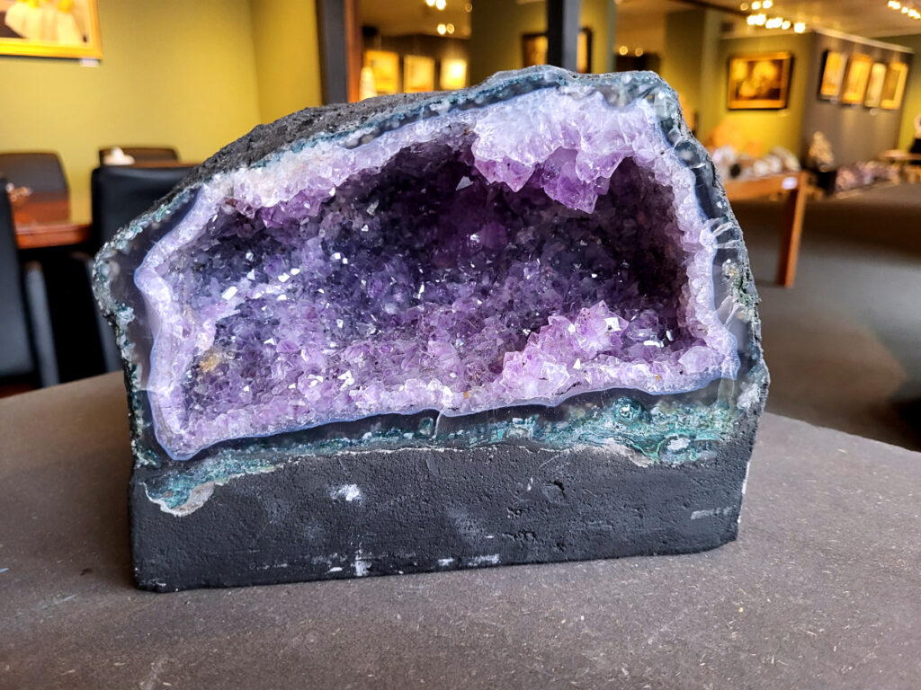 Amethist, Amethist geode met standvlak, Kunstgalerie Arnold, 7,1 kg, 71,- euro, nr. 5

Beschrijving Amethist.
Amethist is een paarse variëteit van kwarts, bekend om zijn kalmerende en spirituele eigenschappen. De kleur varieert van licht lila tot diep violet en wordt veroorzaakt door sporen van ijzer en andere elementen. Amethist wordt al eeuwenlang gewaardeerd, zowel vanwege zijn esthetische schoonheid als zijn vermeende genezende en beschermende krachten. 
Amethist is een kwartskristal, vaak te vinden in geodes (holtes in gesteente) of als afzonderlijke kristallen. 
De kleur varieert van lichtpaars tot diep violet, soms met roze of blauwe tinten. 
De kleurintensiteit kan variëren, waarbij donkerdere amethisten vaak als kwalitatief worden beschouwd. 
Soms vertoont amethist dichroïsme, waarbij je verschillende kleuren kunt zien wanneer je de steen in het licht draait. 

Vindplaatsen:
Amethist wordt gevonden in verschillende delen van de wereld, waaronder Brazilië, Uruguay, Zambia, Madagaskar, Rusland en Canada.
Het wordt vaak gevonden in bergachtige gebieden. 

Gebruik:
Amethist wordt gebruikt in sieraden, decoratieve objecten en als helende steen. 
Het wordt vaak gedragen als hanger, ring of armband, of geplaatst in ruimtes om de energie te zuiveren. 
Amethist wordt ook gebruikt in meditatie en andere spirituele praktijken. 
