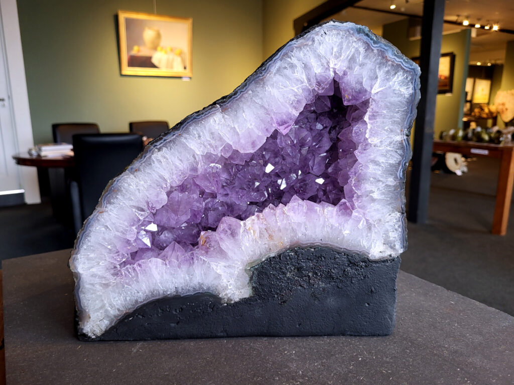 Amethist, Kunstgalerie Arnold, 11,9 kg, 25 cm hoog, 125,- euro, nr. 12

Beschrijving Amethist.
Amethist is een paarse variëteit van kwarts, bekend om zijn kalmerende en spirituele eigenschappen. De kleur varieert van licht lila tot diep violet en wordt veroorzaakt door sporen van ijzer en andere elementen. Amethist wordt al eeuwenlang gewaardeerd, zowel vanwege zijn esthetische schoonheid als zijn vermeende genezende en beschermende krachten. 
Amethist is een kwartskristal, vaak te vinden in geodes (holtes in gesteente) of als afzonderlijke kristallen. 
De kleur varieert van lichtpaars tot diep violet, soms met roze of blauwe tinten. 
De kleurintensiteit kan variëren, waarbij donkerdere amethisten vaak als kwalitatief worden beschouwd. 
Soms vertoont amethist dichroïsme, waarbij je verschillende kleuren kunt zien wanneer je de steen in het licht draait. 

Vindplaatsen:
Amethist wordt gevonden in verschillende delen van de wereld, waaronder Brazilië, Uruguay, Zambia, Madagaskar, Rusland en Canada.
Het wordt vaak gevonden in bergachtige gebieden. 

Gebruik:
Amethist wordt gebruikt in sieraden, decoratieve objecten en als helende steen. 
Het wordt vaak gedragen als hanger, ring of armband, of geplaatst in ruimtes om de energie te zuiveren. 
Amethist wordt ook gebruikt in meditatie en andere spirituele praktijken. 
