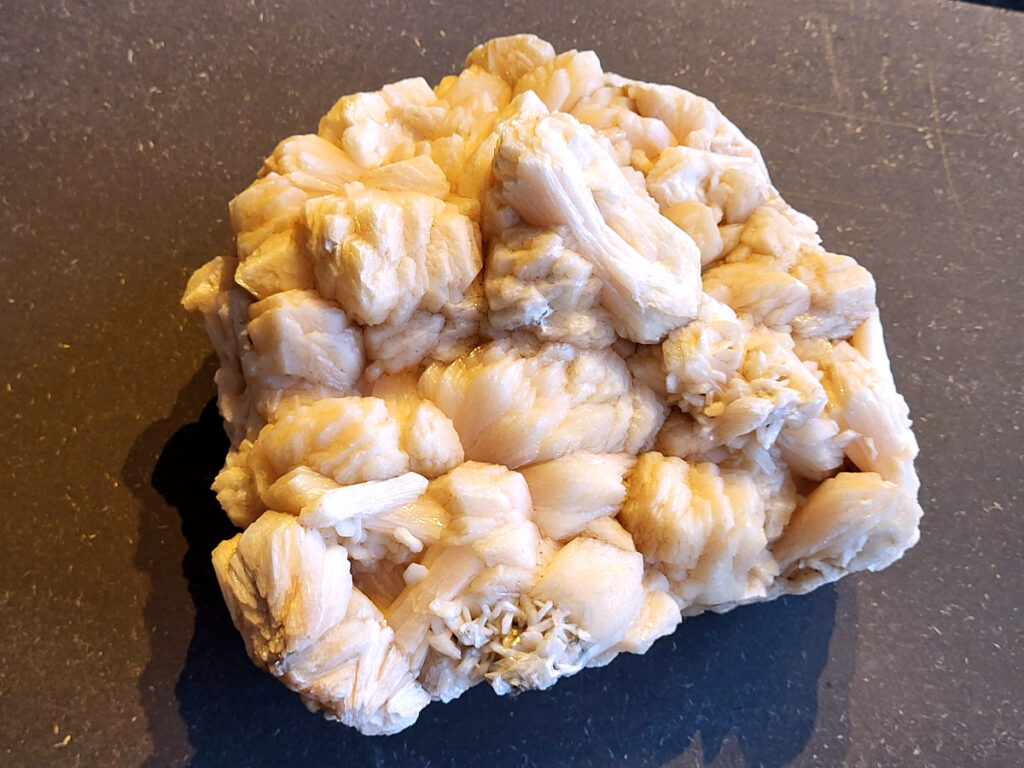 <strong>Stilbiet Cluster, 3,4kg, 67,- euro, nr. 7</strong>

Stilbiet,
<strong>De waaier- of bundelvormige kristallen met een zijdeachtige glans en soms lichtgroen-geelatige gloed zijn typisch voor stilbiet.</strong>
De helder witte tot doorschijnende kubusvormige kristallen aan de basis zijn apofyliet.
De gele of groenige zweem kan komen van een lichte epidoot- of prehniet-insluiting, of een natuurlijke oxidatie aan het oppervlak.
Energetische werking:
<strong>Stilbiet opent het hart en helpt loslaten van zorgen; het is zacht, troostend en bevordert vertrouwen in de loop van het leven.</strong>
Apofyliet versterkt intuïtie, helderheid en spirituele verbinding — een “lichtbrenger” die energie zuivert in een ruimte.
Samen vormen ze een kalmerende, hoogtrillende combinatie die ideaal is voor meditatie, healingruimtes of slaapkamers (voor rust en inzicht).
Zorgtip:
Houd ze uit direct zonlicht (kan verkleuren) en reinig liever met klank, maanlicht of wierook in plaats van water — apofyliet lost iets op bij langdurig vocht.
<strong> Uiterlijk:</strong>
De perzik- tot crème-witte kleur,
De glanzende, waaier- of bloemvormige kristalstructuur,
De zachte, zijdeachtige glans (bijna parelmoerachtig),
al die kenmerken passen heel goed bij stilbiet.
De ronde, lichtgrijze korrelige bol bovenop kan een heulandiet- of chalcedoon-vorming zijn die samen met de stilbiet is gegroeid.
<strong>Energetische betekenis:
Stilbiet hoort bij het hart en het kruinchakra — een zachte, troostende energie die helpt bij het loslaten van oude patronen en het verbinden met hogere leiding.
Ze ondersteunt intuïtie, dromen en zachtheid in relaties — een heel liefdevolle steen om in huis te hebben, vooral bij healing- of slaapruimtes.</strong>