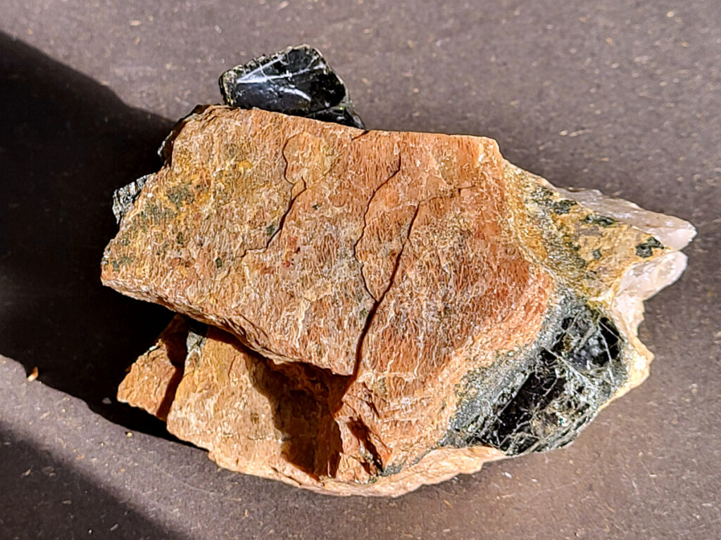 <strong>Zwarte toermalijn (schorl) in een veldspaat-matrix, 0,9 kg, 22,- euro, nr. 2</strong>

Zwarte toermalijn (schorl) in een veldspaat-matrix.
<strong>Zwarte deel: zwarte toermalijn (schorl)
Bruin/roze gesteente eromheen: veldspaat (vaak kaliveldspaat uit pegmatiet)</strong>
<strong>Herkenningspunten</strong>
Zwart kristal met duidelijke lengtestrepen / lijnen
Vaak prismatisch (langwerpig kristal)
<strong>Zit vaak ingegroeid in veldspaat of granietachtige steen</strong>
Komt veel voor in pegmatietgesteente
Dit soort stukken ontstaan diep in de aarde wanneer magma langzaam afkoelt. In pegmatieten kunnen dan grote kristallen groeien, zoals:
Veldspaat, kwarts en toermalijn
Zwarte Toermalijn in Veldspaat
<strong>Zwarte toermalijn in veldspaat combineert de aardende en beschermende eigenschappen van zwarte toermalijn met de harmoniserende energieën van veldspaat.</strong> Deze unieke steen wordt gevormd wanneer zwarte toermalijnkristallen op natuurlijke wijze zijn ingebed in de veldspaatmatrix, waardoor een opvallend contrast ontstaat tussen de donkere toermalijn en de lichtere veldspaat.
<strong>Zwarte toermalijn staat bekend om zijn vermogen om negatieve energieën te absorberen en psychische bescherming te bieden.</strong> Het fungeert als een schild, buigt schadelijke energieën af en bevordert een gevoel van veiligheid en zekerheid. Met zijn aardende eigenschappen helpt zwarte toermalijn de drager te verankeren in de aarde, waardoor een gevoel van stabiliteit en evenwicht wordt bevorderd.
<strong>Veldspaat daarentegen is een steen van harmonie en integratie. Het bevordert een gevoel van eenheid en samenwerking, zowel binnen jezelf als in relaties met anderen. Veldspaat staat ook bekend om zijn kalmerende en verzachtende eigenschappen, waardoor stress en angst worden verlicht.</strong>
Samen creëert zwarte toermalijn in veldspaat een krachtige synergie van energieën. Het dient als een krachtig hulpmiddel voor spirituele bescherming en aarding, terwijl het ook innerlijke harmonie en emotioneel evenwicht bevordert. <strong>Deze steen kan bijzonder nuttig zijn voor degenen die een gevoel van stabiliteit en veiligheid in hun leven willen creëren, terwijl ze ook een diepere verbinding met de aarde en het spirituele rijk bevorderen.</strong>