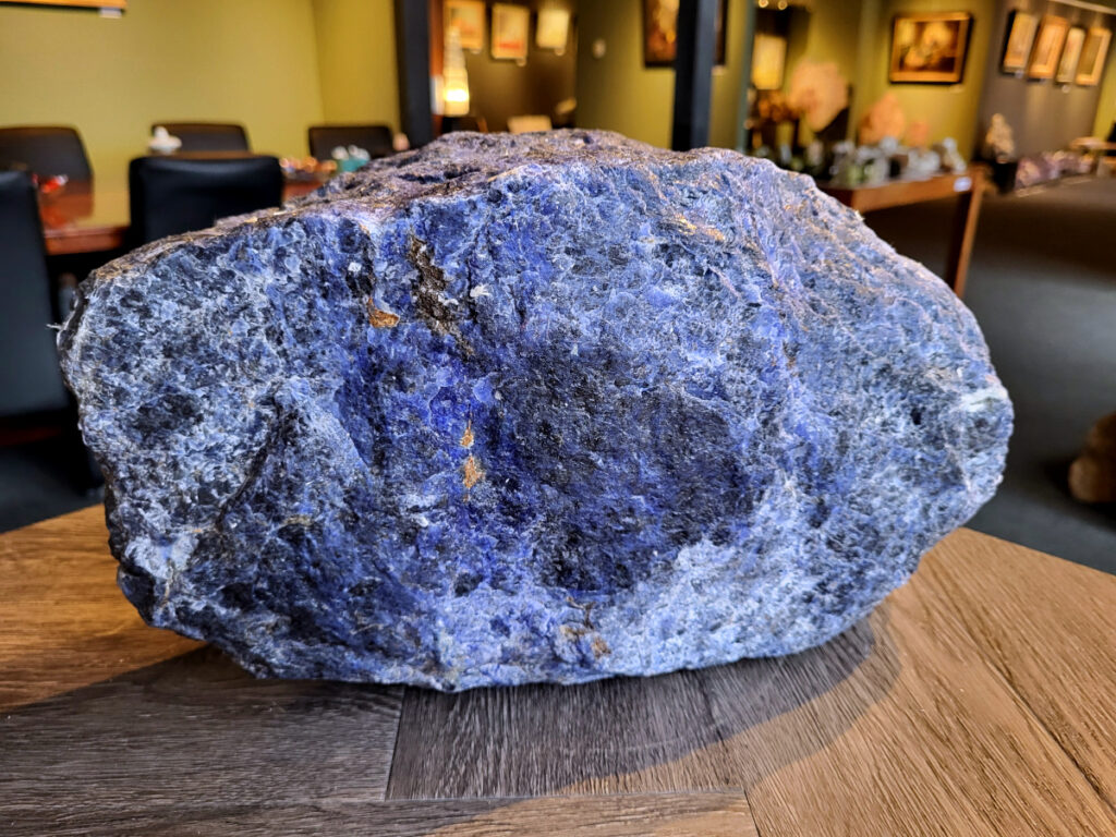 Sodaliet, ruw, 13 kg, 35cm doorsnee, 90,- euro, nr. 1

Sodaliet, beschrijving,
Sodaliet is een diepblauwe edelsteen en mineraal, bekend om zijn helderheid, spirituele betekenis en helende eigenschappen.
Sodaliet is een natrium-aluminiumsilicaat dat behoort tot de veldspaatmineralen en vaak witte aders of vlekken bevat, hoewel de mooiste kwaliteit inktblauw en zonder aders is (Sodaliet kan ook groen, rood of roze zijn afhankelijk van de samenstelling) . De steen heeft een hardheid van ongeveer 5,5–6 op de schaal van Mohs, een glasachtige tot matte glans en een gemiddelde dichtheid van 2,3–2,4 g/cm³. De naam sodaliet is afgeleid van het Engelse sodium (natrium) en het Griekse lithos (steen).  
Geschiedenis en gebruik
Sodaliet werd voor het eerst ontdekt in Zuid-Amerika, met name in Chili, en later ook in Brazilië en Uruguay. Het werd al snel populair als edelsteen en decoratief materiaal, gebruikt in sieraden, beeldjes, schalen en zelfs als blauwe verf of oogmake-up in de oudheid  Oude culturen zoals de Egyptenaren, Grieken en Romeinen waardeerden sodaliet om zijn beschermende en concentratie bevorderende eigenschappen . 
Spirituele en helende eigenschappen
Sodaliet staat bekend als de Steen van de Denker en wordt geassocieerd met rationaliteit, objectiviteit, intuïtie en innerlijke waarheid. Het helpt bij het structureren van gedachten, helder communiceren en zelfvertrouwen, en bevordert rust en focus tijdens werk, studie of meditatie  Spiritueel opent sodaliet het derde oogchakra, versterkt intuïtie en ondersteunt inzichtgevende meditaties. De steen wordt ook gebruikt om negatieve energie af te weren en harmonie in groepen te bevorderen. 
Fysieke voordelen
Fysiek kan sodaliet helpen bij keel-, hals- en stembandproblemen, het verlagen van bloeddruk, het ontspannen van nek en schouders, en het ondersteunen van het beenderstelsel en herstel van pezen  Dankzij het natriumgehalte heeft sodaliet ook een desinfecterende werking. 
Toepassingen
Sodaliet wordt vaak gebruikt in sieraden zoals kettingen, ringen en oorbellen, maar ook voor decoratieve objecten, houtsnijwerk en wandbedekking Het past goed bij de sterrenbeelden Boogschutter en Maagd, en kan helpen bij het vinden van focus, richting en zelfvertrouwen. 
Al met al is sodaliet een veelzijdige steen die zowel mentaal, emotioneel, fysiek als spiritueel ondersteuning biedt en geliefd is om zijn schoonheid en krachtige energetische eigenschappen.