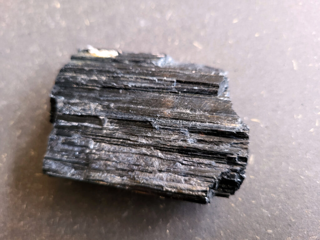 <strong>Toermalijn, zwarte toermalijn, 5,50 euro, nr. 5

Zwarte toermalijn, beschrijving
Zwarte toermalijn (schorl) in een veldspaat-matrix.
Zwarte deel: zwarte toermalijn (schorl)
Bruin/roze gesteente eromheen: veldspaat (vaak kaliveldspaat uit pegmatiet)
Herkenningspunten
Zwart kristal met duidelijke lengtestrepen / lijnen
Vaak prismatisch (langwerpig kristal)
Zit vaak ingegroeid in veldspaat of granietachtige steen
Komt veel voor in pegmatietgesteente
Dit soort stukken ontstaan diep in de aarde wanneer magma langzaam afkoelt. In pegmatieten kunnen dan grote kristallen groeien, zoals:
Veldspaat, kwarts en toermalijn
Zwarte Toermalijn in Veldspaat
Zwarte toermalijn in veldspaat combineert de aardende en beschermende eigenschappen van zwarte toermalijn met de harmoniserende energieën van veldspaat. Deze unieke steen wordt gevormd wanneer zwarte toermalijnkristallen op natuurlijke wijze zijn ingebed in de veldspaatmatrix, waardoor een opvallend contrast ontstaat tussen de donkere toermalijn en de lichtere veldspaat.
Zwarte toermalijn staat bekend om zijn vermogen om negatieve energieën te absorberen en psychische bescherming te bieden. Het fungeert als een schild, buigt schadelijke energieën af en bevordert een gevoel van veiligheid en zekerheid. Met zijn aardende eigenschappen helpt zwarte toermalijn de drager te verankeren in de aarde, waardoor een gevoel van stabiliteit en evenwicht wordt bevorderd.
Veldspaat daarentegen is een steen van harmonie en integratie. Het bevordert een gevoel van eenheid en samenwerking, zowel binnen jezelf als in relaties met anderen. Veldspaat staat ook bekend om zijn kalmerende en verzachtende eigenschappen, waardoor stress en angst worden verlicht.
Samen creëert zwarte toermalijn in veldspaat een krachtige synergie van energieën. Het dient als een krachtig hulpmiddel voor spirituele bescherming en aarding, terwijl het ook innerlijke harmonie en emotioneel evenwicht bevordert. Deze steen kan bijzonder nuttig zijn voor degenen die een gevoel van stabiliteit en veiligheid in hun leven willen creëren, terwijl ze ook een diepere verbinding met de aarde en het spirituele rijk bevorderen.</strong>