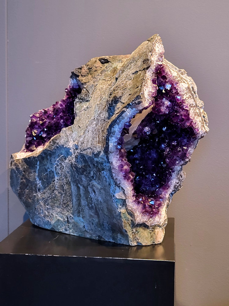 Amethist, cluster, geode, 36cm hoog en 42cm breed, ruim 25 kg, 1650,- euro, nr. 1

Beschrijving Amethist.
Amethist is een paarse variëteit van kwarts, bekend om zijn kalmerende en spirituele eigenschappen. De kleur varieert van licht lila tot diep violet en wordt veroorzaakt door sporen van ijzer en andere elementen. Amethist wordt al eeuwenlang gewaardeerd, zowel vanwege zijn esthetische schoonheid als zijn vermeende genezende en beschermende krachten.
Amethist is een kwartskristal, vaak te vinden in geodes (holtes in gesteente) of als afzonderlijke kristallen.
De kleur varieert van lichtpaars tot diep violet, soms met roze of blauwe tinten.
De kleurintensiteit kan variëren, waarbij donkerdere amethisten vaak als kwalitatief worden beschouwd.
Soms vertoont amethist dichroïsme, waarbij je verschillende kleuren kunt zien wanneer je de steen in het licht draait.

Vindplaatsen:
Amethist wordt gevonden in verschillende delen van de wereld, waaronder Brazilië, Uruguay, Zambia, Madagaskar, Rusland en Canada.
Het wordt vaak gevonden in bergachtige gebieden.

Gebruik:
Amethist wordt gebruikt in sieraden, decoratieve objecten en als helende steen.
Het wordt vaak gedragen als hanger, ring of armband, of geplaatst in ruimtes om de energie te zuiveren.
Amethist wordt ook gebruikt in meditatie en andere spirituele praktijken.