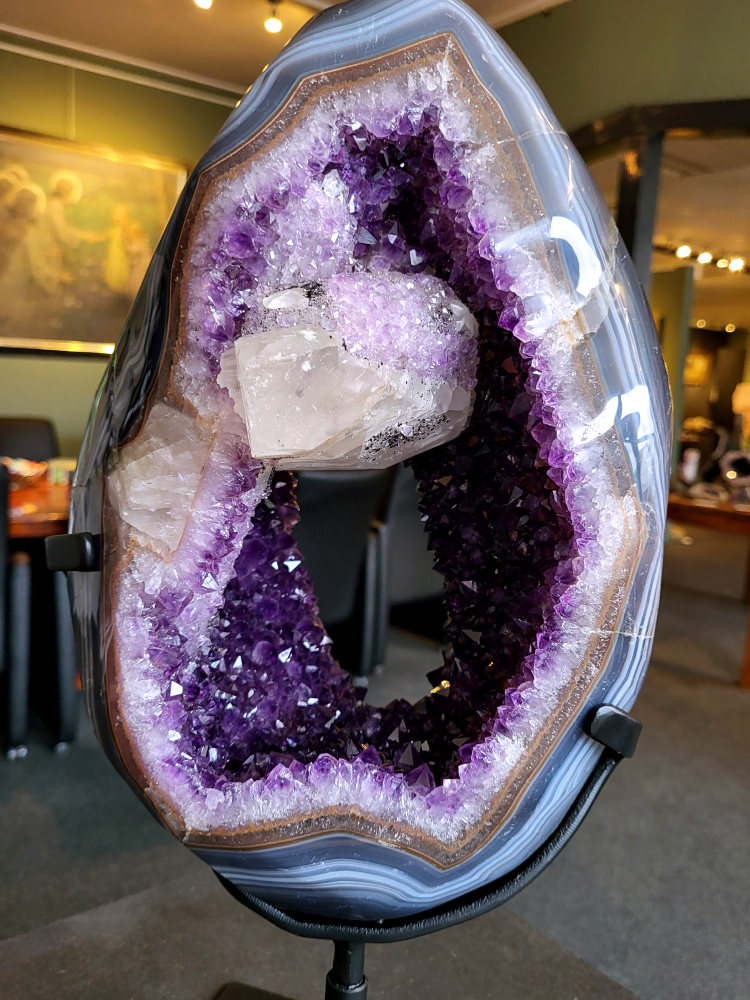 <strong>Amethist Geode met calciet en agaat, 12,7kg, 48cm hoog, 25cm doorsnee, 1350,- euro, nr. 42

Beschrijving Amethist.
Amethist is een paarse variëteit van kwarts, bekend om zijn kalmerende en spirituele eigenschappen. De kleur varieert van licht lila tot diep violet en wordt veroorzaakt door sporen van ijzer en andere elementen. Amethist wordt al eeuwenlang gewaardeerd, zowel vanwege zijn esthetische schoonheid als zijn vermeende genezende en beschermende krachten.
Amethist is een kwartskristal, vaak te vinden in geodes (holtes in gesteente) of als afzonderlijke kristallen.
De kleur varieert van lichtpaars tot diep violet, soms met roze of blauwe tinten.
De kleurintensiteit kan variëren, waarbij donkerdere amethisten vaak als kwalitatief worden beschouwd.
Soms vertoont amethist dichroïsme, waarbij je verschillende kleuren kunt zien wanneer je de steen in het licht draait.

Vindplaatsen:
Amethist wordt gevonden in verschillende delen van de wereld, waaronder Brazilië, Uruguay, Zambia, Madagaskar, Rusland en Canada.
Het wordt vaak gevonden in bergachtige gebieden.

Gebruik:
Amethist wordt gebruikt in sieraden, decoratieve objecten en als helende steen.
Het wordt vaak gedragen als hanger, ring of armband, of geplaatst in ruimtes om de energie te zuiveren.
Amethist wordt ook gebruikt in meditatie en andere spirituele praktijken.</strong>
