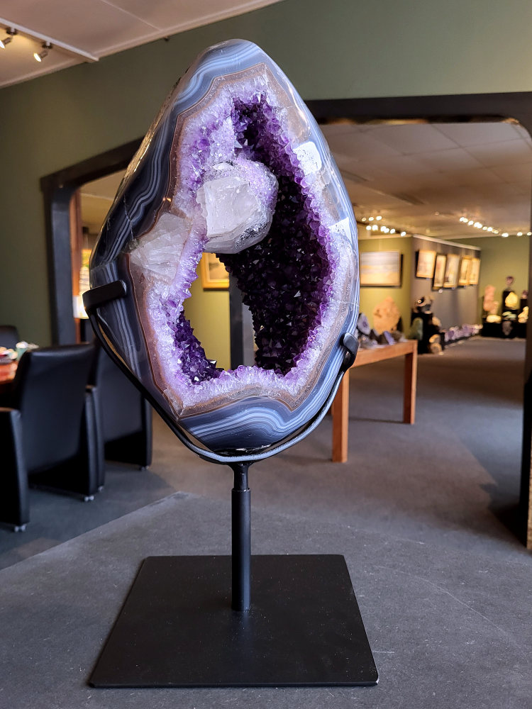 <strong>Amethist Geode met calciet en agaat, 12,7kg, 48cm hoog, 25cm doorsnee, 1350,- euro, nr. 43

Beschrijving Amethist.
Amethist is een paarse variëteit van kwarts, bekend om zijn kalmerende en spirituele eigenschappen. De kleur varieert van licht lila tot diep violet en wordt veroorzaakt door sporen van ijzer en andere elementen. Amethist wordt al eeuwenlang gewaardeerd, zowel vanwege zijn esthetische schoonheid als zijn vermeende genezende en beschermende krachten.
Amethist is een kwartskristal, vaak te vinden in geodes (holtes in gesteente) of als afzonderlijke kristallen.
De kleur varieert van lichtpaars tot diep violet, soms met roze of blauwe tinten.
De kleurintensiteit kan variëren, waarbij donkerdere amethisten vaak als kwalitatief worden beschouwd.
Soms vertoont amethist dichroïsme, waarbij je verschillende kleuren kunt zien wanneer je de steen in het licht draait.

Vindplaatsen:
Amethist wordt gevonden in verschillende delen van de wereld, waaronder Brazilië, Uruguay, Zambia, Madagaskar, Rusland en Canada.
Het wordt vaak gevonden in bergachtige gebieden.

Gebruik:
Amethist wordt gebruikt in sieraden, decoratieve objecten en als helende steen.
Het wordt vaak gedragen als hanger, ring of armband, of geplaatst in ruimtes om de energie te zuiveren.
Amethist wordt ook gebruikt in meditatie en andere spirituele praktijken.</strong>