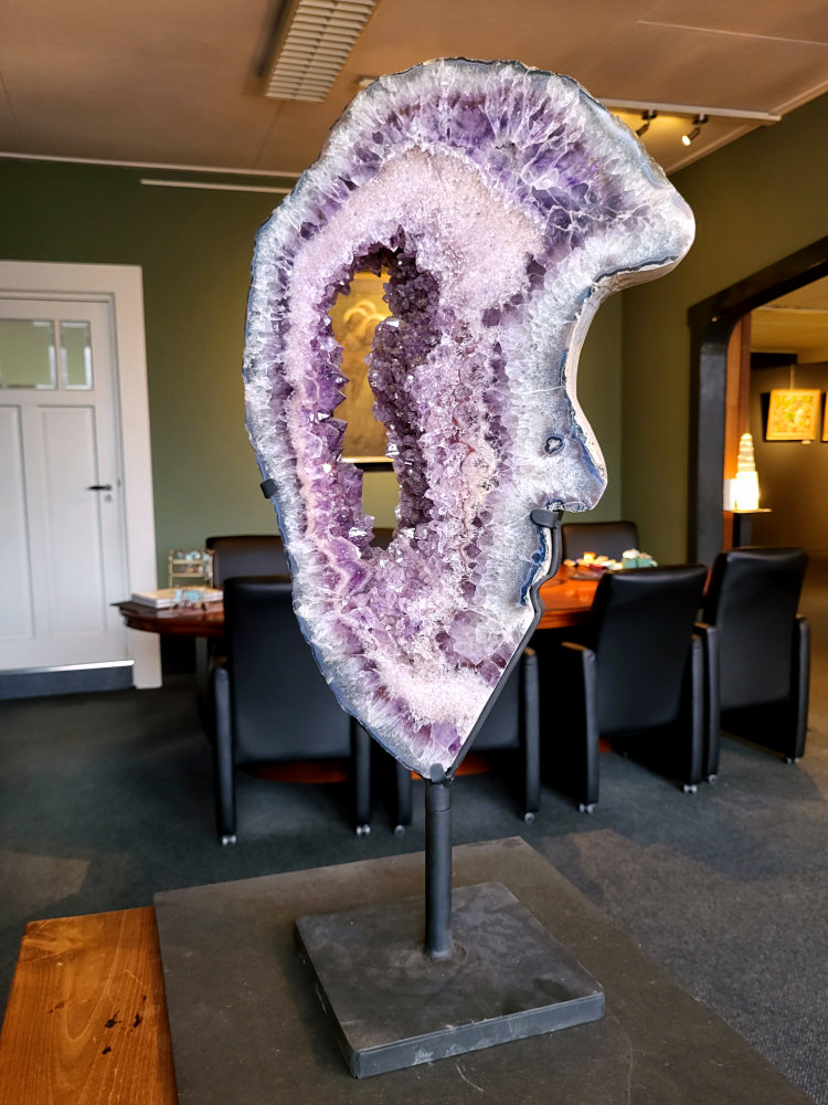 <strong>Amethist met Chalcedoon op standaard,15,7kg, 75cm hoog, 850,- euro, nr. 44

Beschrijving Amethist.
Amethist is een paarse variëteit van kwarts, bekend om zijn kalmerende en spirituele eigenschappen. De kleur varieert van licht lila tot diep violet en wordt veroorzaakt door sporen van ijzer en andere elementen. Amethist wordt al eeuwenlang gewaardeerd, zowel vanwege zijn esthetische schoonheid als zijn vermeende genezende en beschermende krachten.
Amethist is een kwartskristal, vaak te vinden in geodes (holtes in gesteente) of als afzonderlijke kristallen.
De kleur varieert van lichtpaars tot diep violet, soms met roze of blauwe tinten.
De kleurintensiteit kan variëren, waarbij donkerdere amethisten vaak als kwalitatief worden beschouwd.
Soms vertoont amethist dichroïsme, waarbij je verschillende kleuren kunt zien wanneer je de steen in het licht draait.

Vindplaatsen:
Amethist wordt gevonden in verschillende delen van de wereld, waaronder Brazilië, Uruguay, Zambia, Madagaskar, Rusland en Canada.
Het wordt vaak gevonden in bergachtige gebieden.

Gebruik:
Amethist wordt gebruikt in sieraden, decoratieve objecten en als helende steen.
Het wordt vaak gedragen als hanger, ring of armband, of geplaatst in ruimtes om de energie te zuiveren.
Amethist wordt ook gebruikt in meditatie en andere spirituele praktijken.</strong>